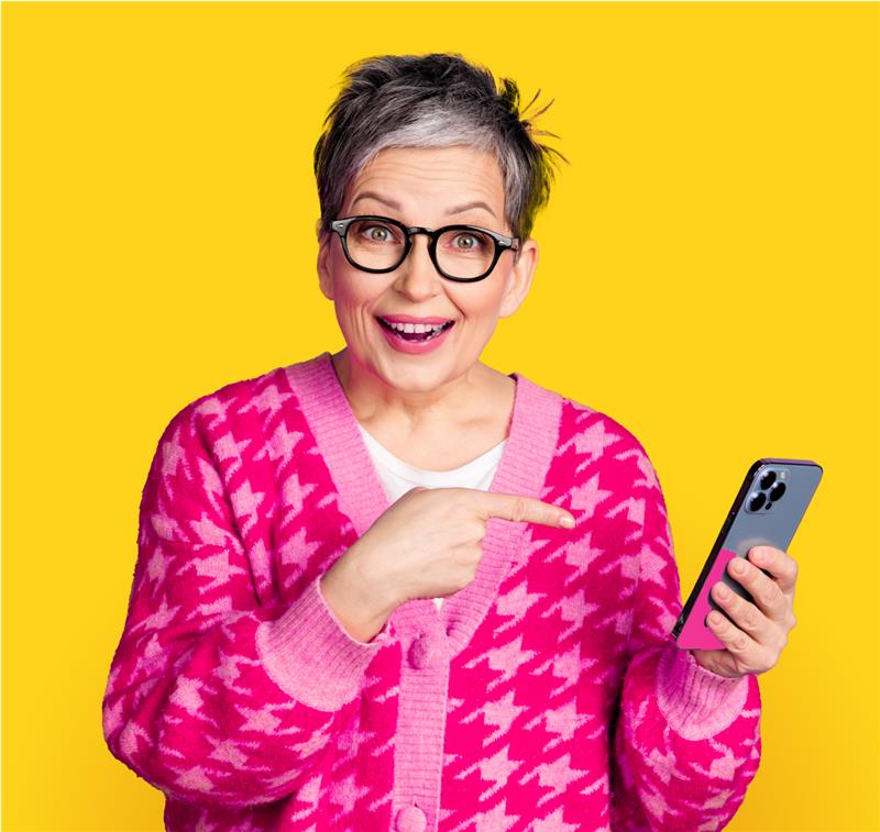 Starsza kobieta w okularach i różowym swetrze uśmiecha się i wskazuje palcem na trzymany w ręku smartfon. Tło jest jednolicie żółte.