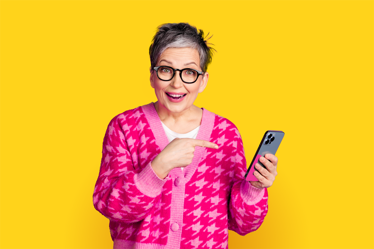 Starsza kobieta w okularach i różowym swetrze uśmiecha się i wskazuje palcem na trzymany w ręku smartfon. Tło jest jednolicie żółte.
