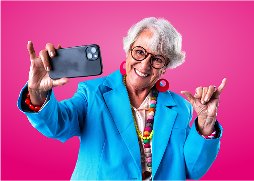Starsza kobieta w jasnoniebieskiej marynarce i kolorowej biżuterii robi selfie smartfonem na tle intensywnie różowego tła. Uśmiecha się, nosi okulary i pokazuje gest hang loose drugą ręką.