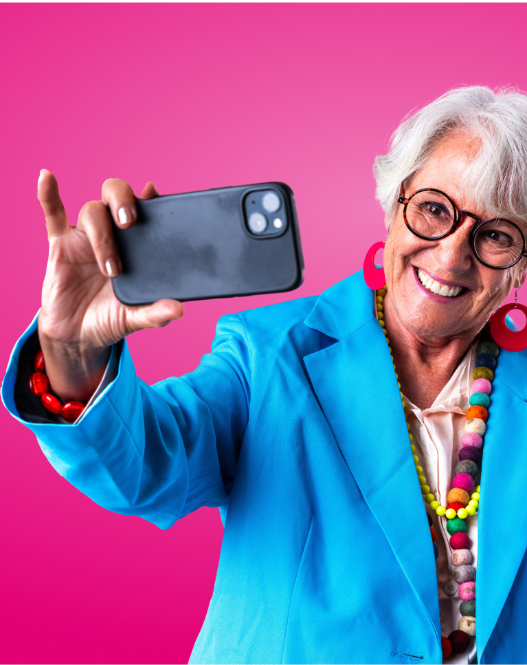Starsza kobieta w jasnoniebieskiej marynarce i kolorowej biżuterii robi selfie smartfonem na tle intensywnie różowego tła. Uśmiecha się, nosi okulary i pokazuje gest hang loose drugą ręką.